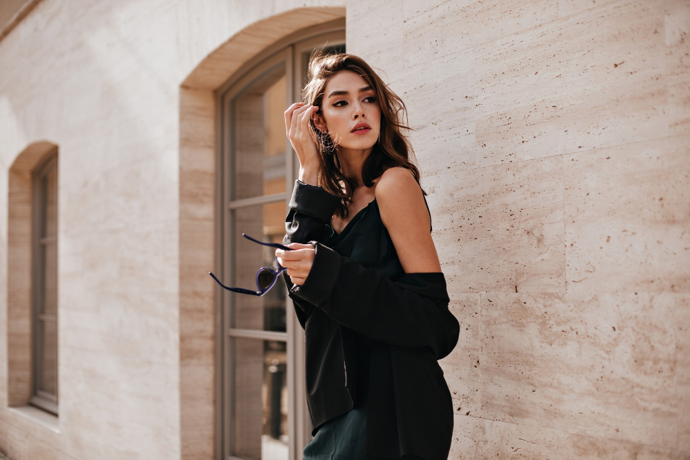 cute-young-girl-with-dark-wavy-hairstyle-bright-makeup-silk-dress-black-jacket-holding-sunglasses-hands-looking-away-against-beige-building-wall